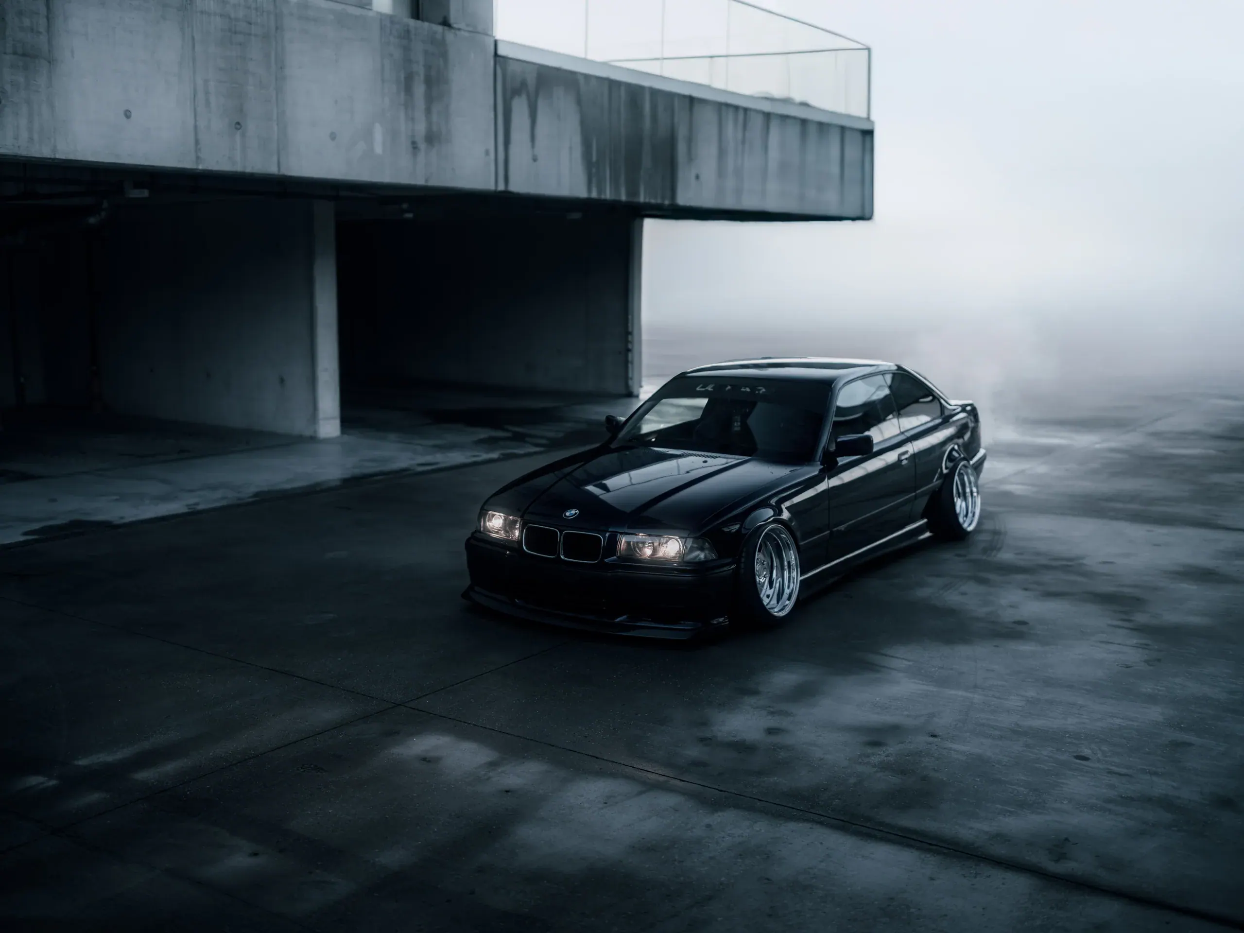 BMW E36 ustawione pod betonowym okapem wielopoziomowego parkingu.