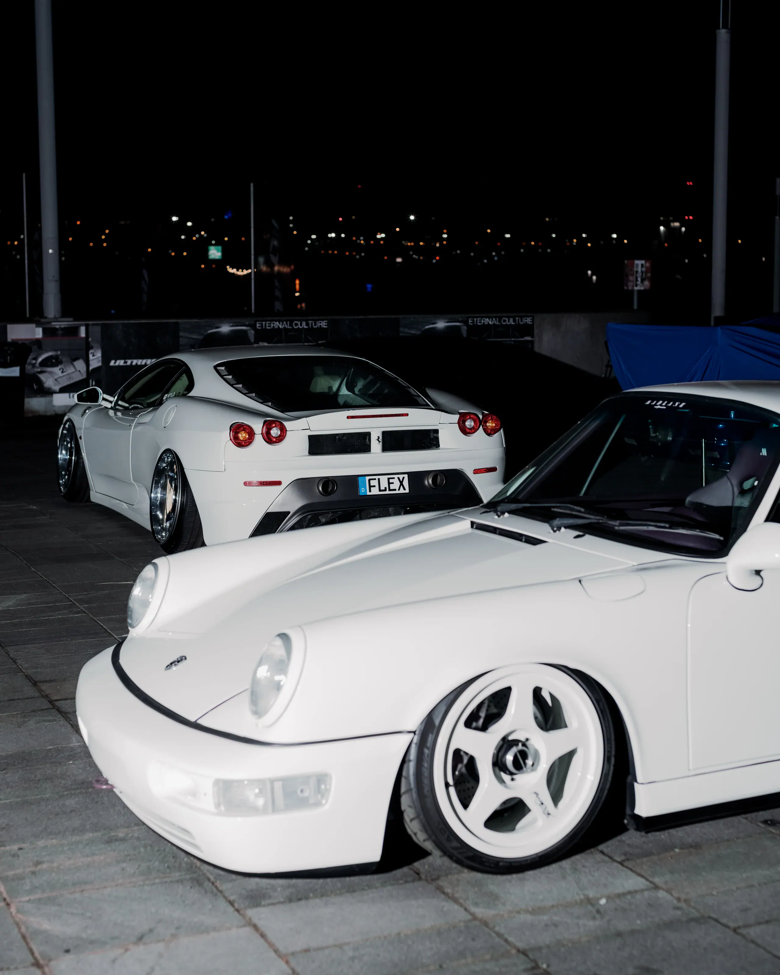Przedni narożnik białego Porsche na tle nocnego zlotu samochodowego.