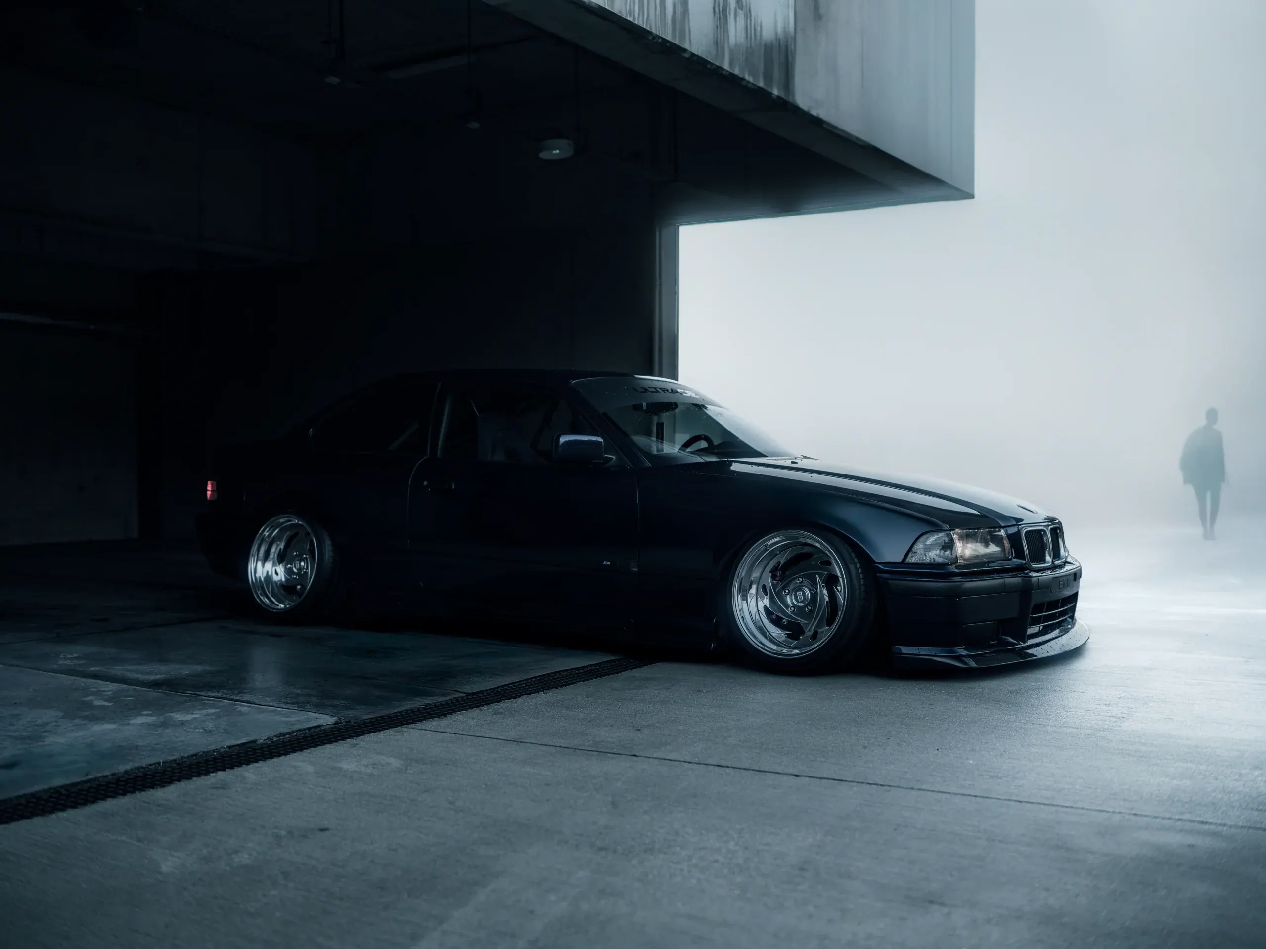 Ciemne BMW E36 widziane z boku pod surową, betonową konstrukcją parkingu.